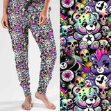 Full length jogger pants featuring colourful punk-inspired teddy bears, rainbow skulls, bats, spiderwebs and neon alternative artwork on a black background with bright pink, purple, green and blue accents.