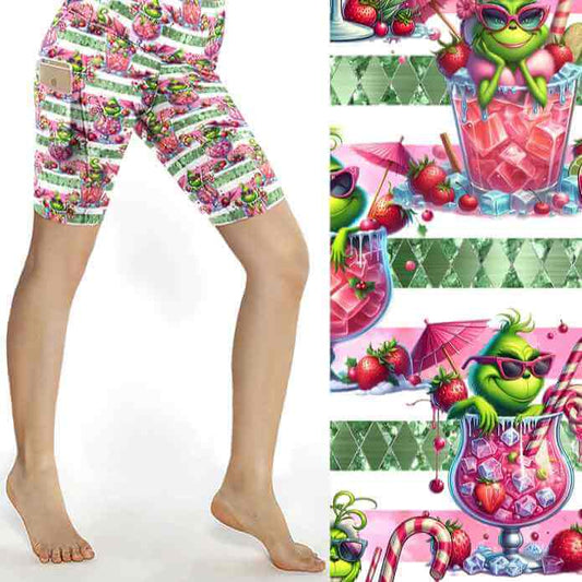 Christmas sports shorts featuring a green Grinch-like character lounging in pink cocktail glasses on a pink and white striped background with fruit and candy canes by Nikki Whoops Boutique.