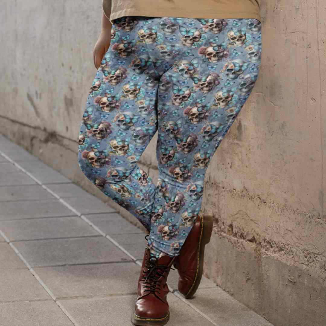 Person wearing light blue leggings with a pattern of skulls, flowers, and butterflies, leaning against a concrete wall. The outfit is paired with brown lace-up boots and a loose beige top, creating a casual and stylish look. The leggings' design features pastel florals and blue butterflies on skulls, adding a whimsical touch.
