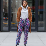 Woman wearing a brown cardigan, white hoodie, Full length jogger pants featuring colourful cosmic cats with bright blue, pink and purple fur, yellow eyes and mischievous smiles standing on a city street.