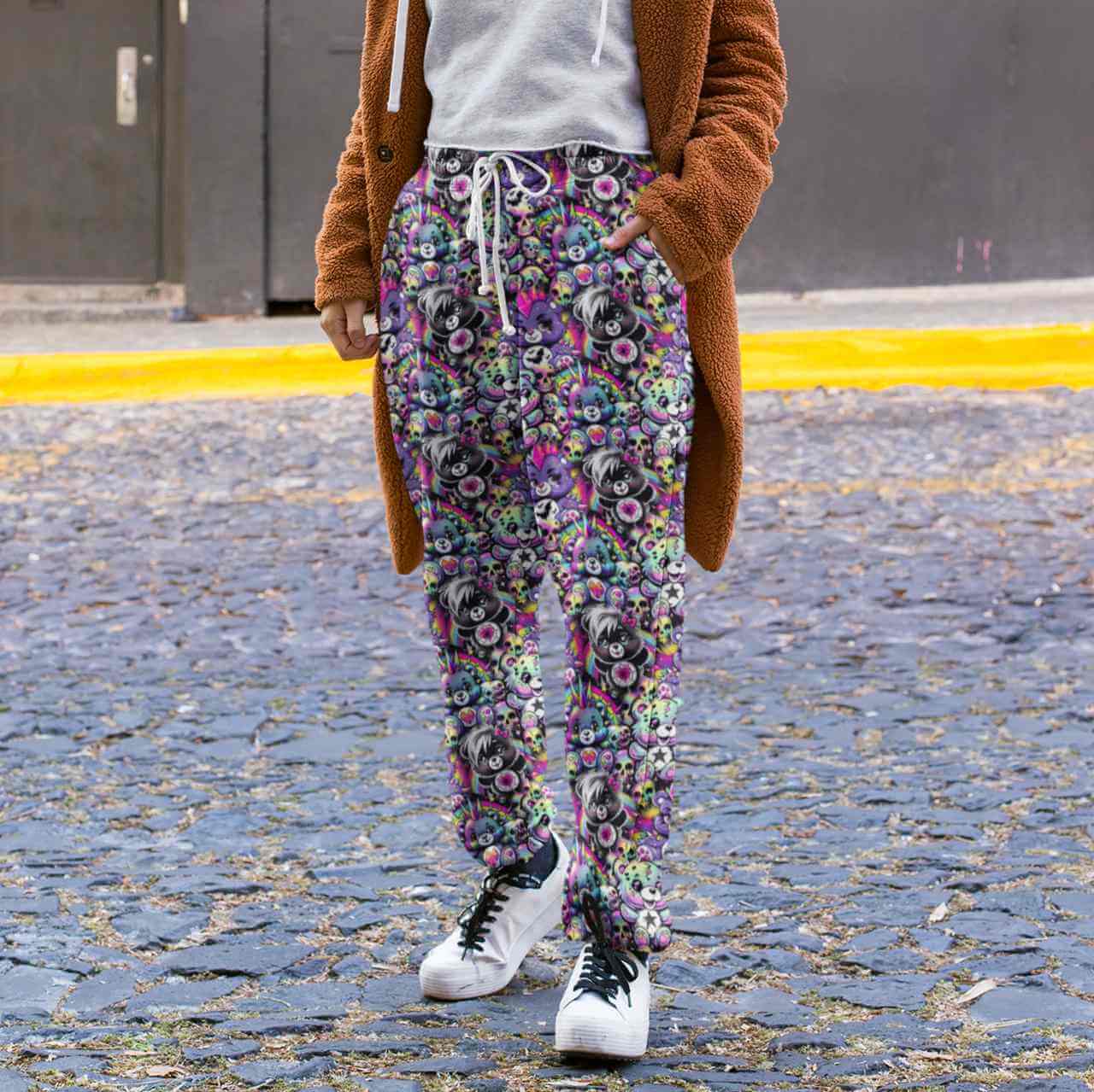 Person wearing Full length jogger pants featuring colourful punk-inspired teddy bears, rainbow skulls, bats, spiderwebs and neon alternative artwork on a black background with bright pink, purple, green and blue accents and a brown coat on a street.