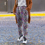 Person wearing Full length jogger pants featuring colourful punk-inspired teddy bears, rainbow skulls, bats, spiderwebs and neon alternative artwork on a black background with bright pink, purple, green and blue accents and a brown coat on a street.