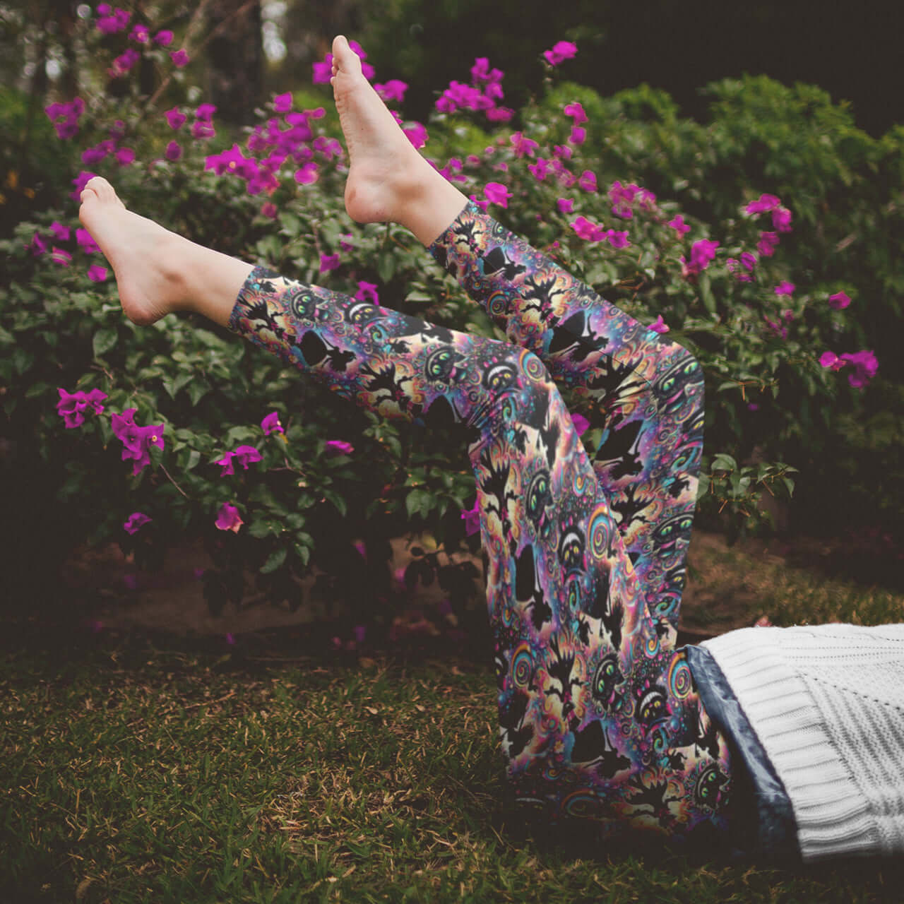 Person wearing colorful patterned pants with feet raised in the air against a background of greenery and pink flowers.