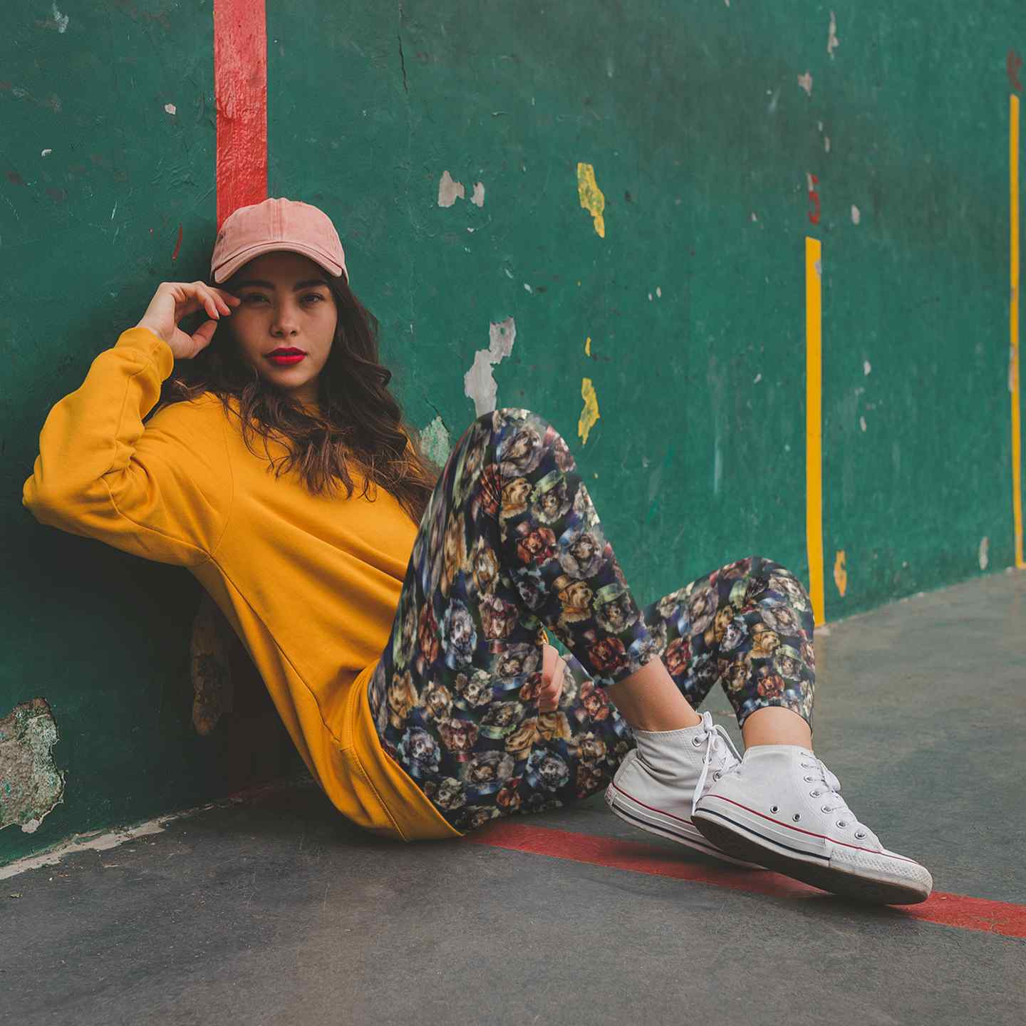 Woman sitting on a basketball court wearing a yellow sweatshirt, floral pants, and white sneakers.