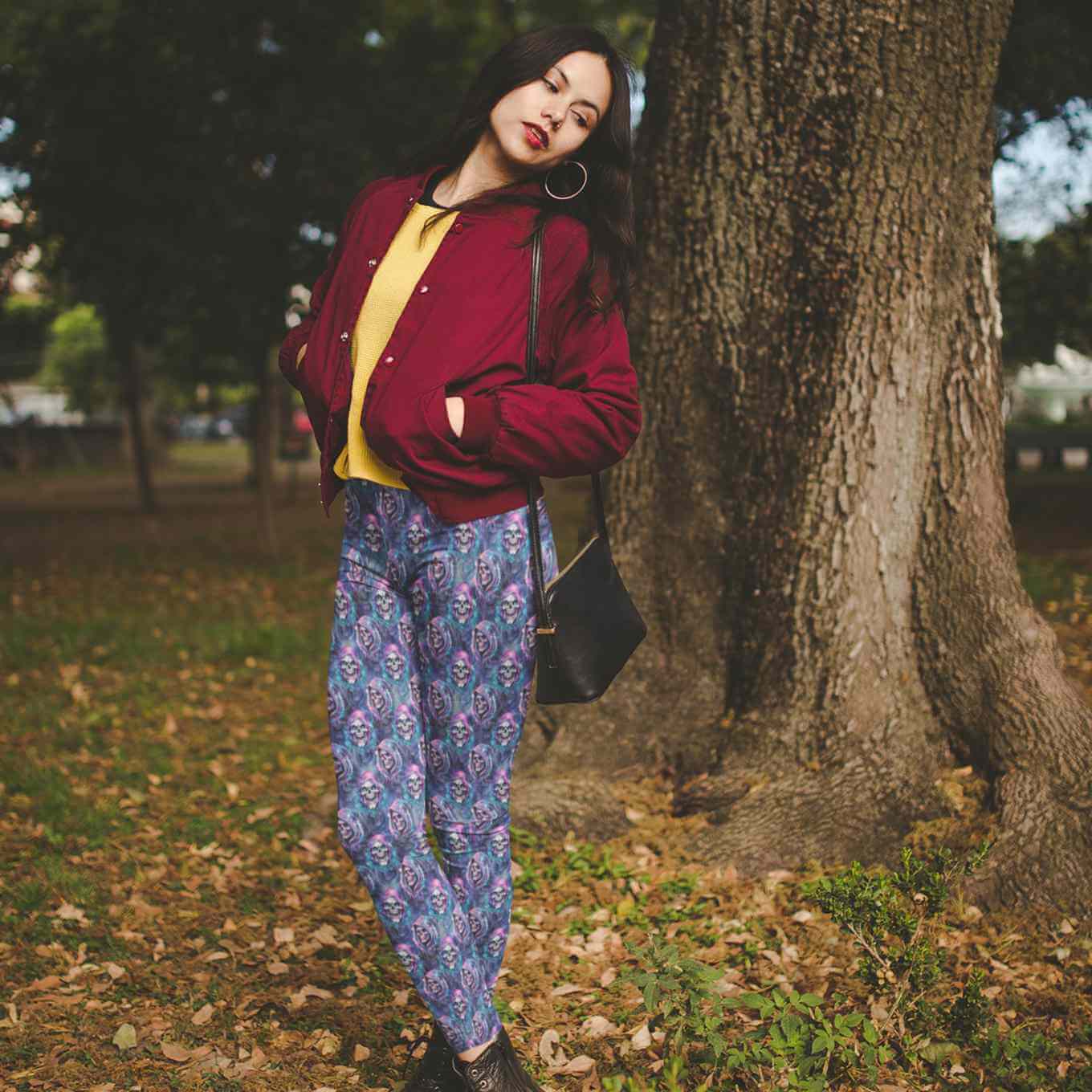 Woman in a red jacket and leggings with a colorful sparkly Grim reaper skull design on blue background standing next to a tree in a park.
