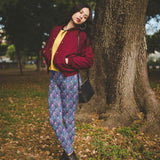 Woman in a red jacket and leggings with a colorful sparkly Grim reaper skull design on blue background standing next to a tree in a park.