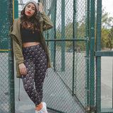 Woman in green jacket and patterned pants standing in front of a chain-link fence.