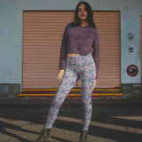 Woman wearing a purple sweater and leggings featuring pattern of skeletons on pink flamingo floats with umbrellas against a blue marble background standing in front of a closed shutter.