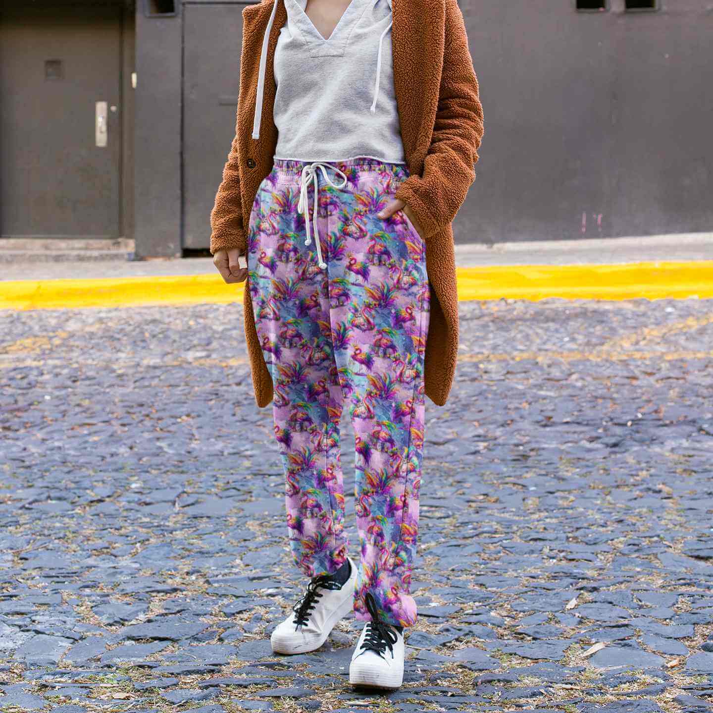 lady on a cobblestone street wearing Full-length jogger pants featuring vibrant pink flamingos dressed in colourful Mardi Gras-style feathers and accessories. The background is a swirling galaxy of purples, pinks and blues with stars and glittery textures.