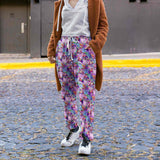lady on a cobblestone street wearing Full-length jogger pants featuring vibrant pink flamingos dressed in colourful Mardi Gras-style feathers and accessories. The background is a swirling galaxy of purples, pinks and blues with stars and glittery textures.