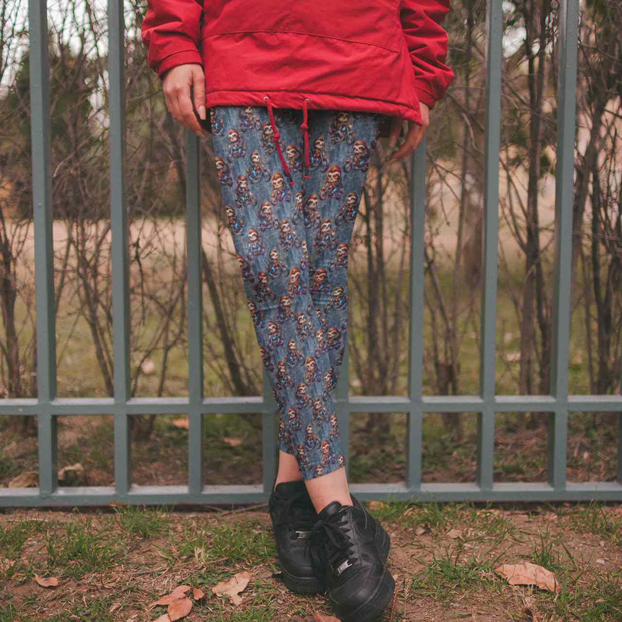 Person wearing a red jacket,  sloths riding motorcycles on a denim background, and black boots standing in front of a metal fence.