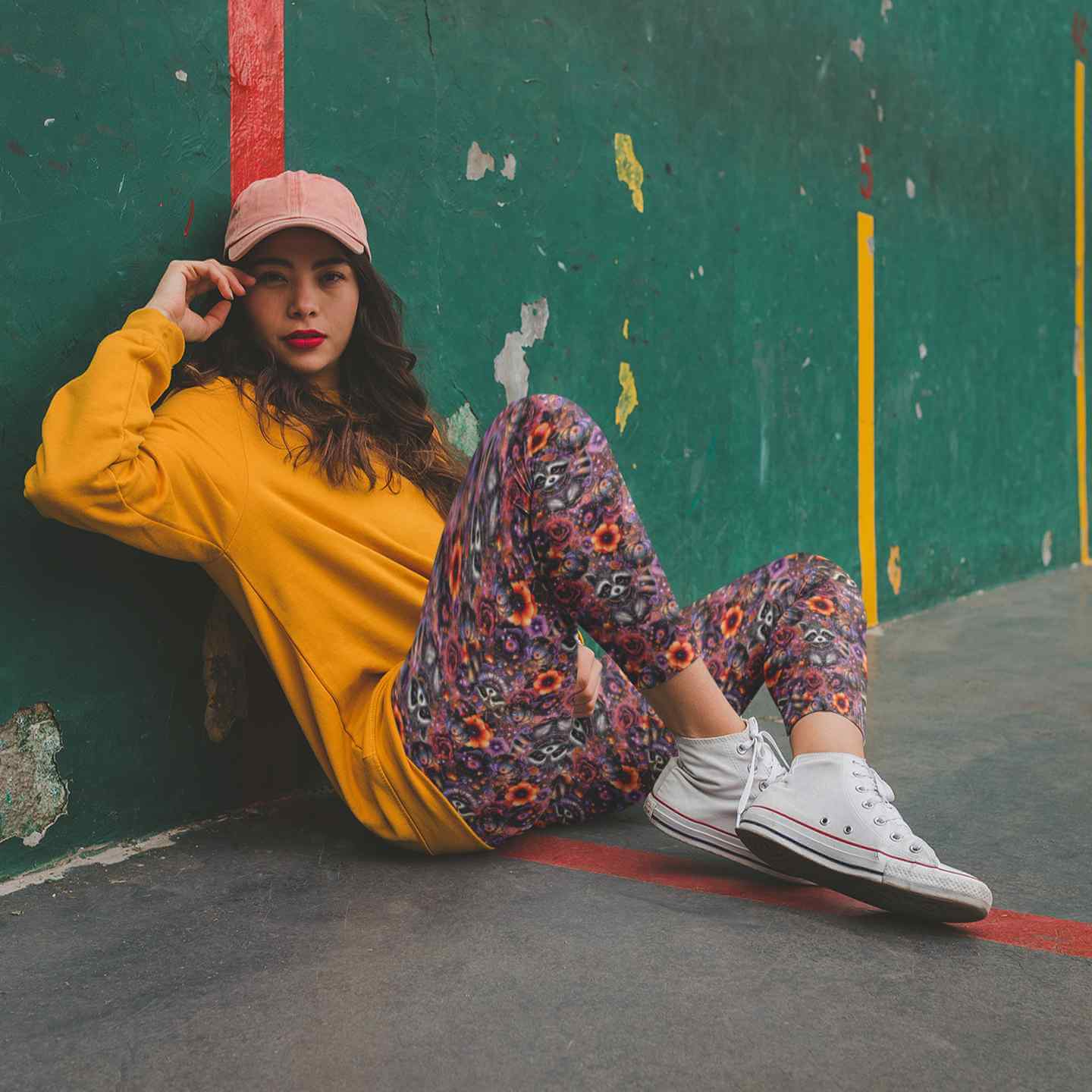 Woman sitting against a green wall wearing a yellow sweatshirt, raccoon leggings, and white sneakers.
