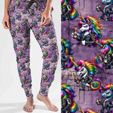 mockup of Full-length jogger pants with a bold print featuring punk-style unicorns with rainbow manes, leather jackets, studs and beer bottles. The background is a grungy purple brick wall for added edge.