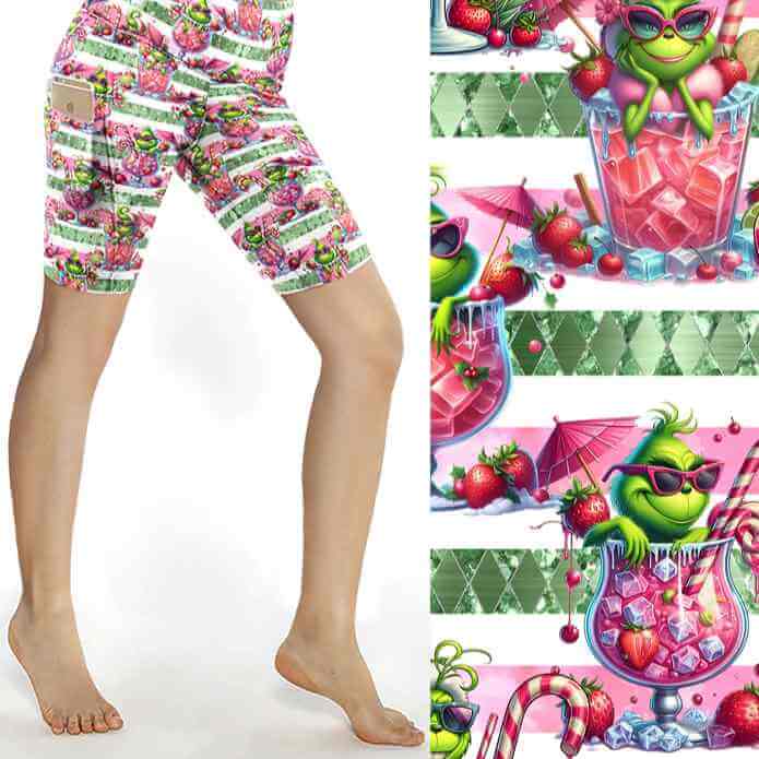 Christmas sports shorts featuring a green Grinch-like character lounging in pink cocktail glasses on a pink and white striped background with fruit and candy canes by Nikki Whoops Boutique.