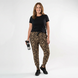 A woman modeling leopard-print jogger pants with a black t-shirt and black sneakers, holding a water bottle against a plain white studio background.