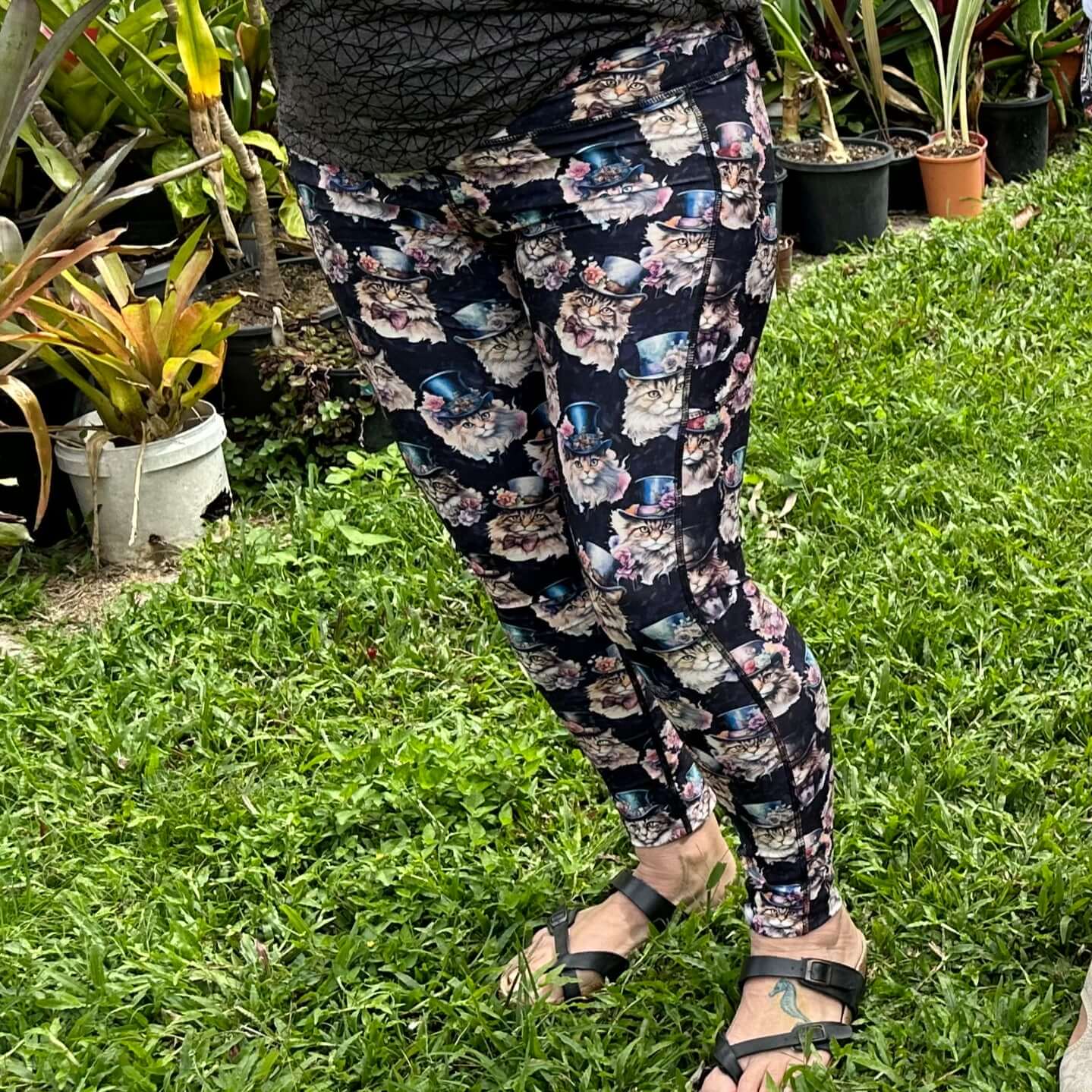 Person wearing leggings featuring a whimsical Victorian-inspired cat print, with a wide waistband and deep side pockets, standing on grass with plants in the background