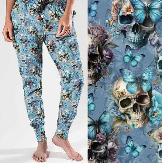 Mockup of full-length jogger pants in a baby blue colour featuring an all-over print of realistic skulls, blue butterflies and pastel flowers. The design blends dark gothic elements with soft, feminine details.