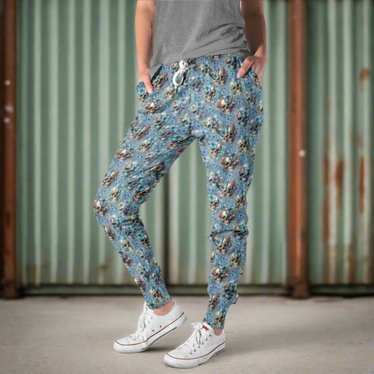 Mockup of lady wearing full-length jogger pants in a baby blue colour featuring an all-over print of realistic skulls, blue butterflies and pastel flowers. The design blends dark gothic elements with soft, feminine details.