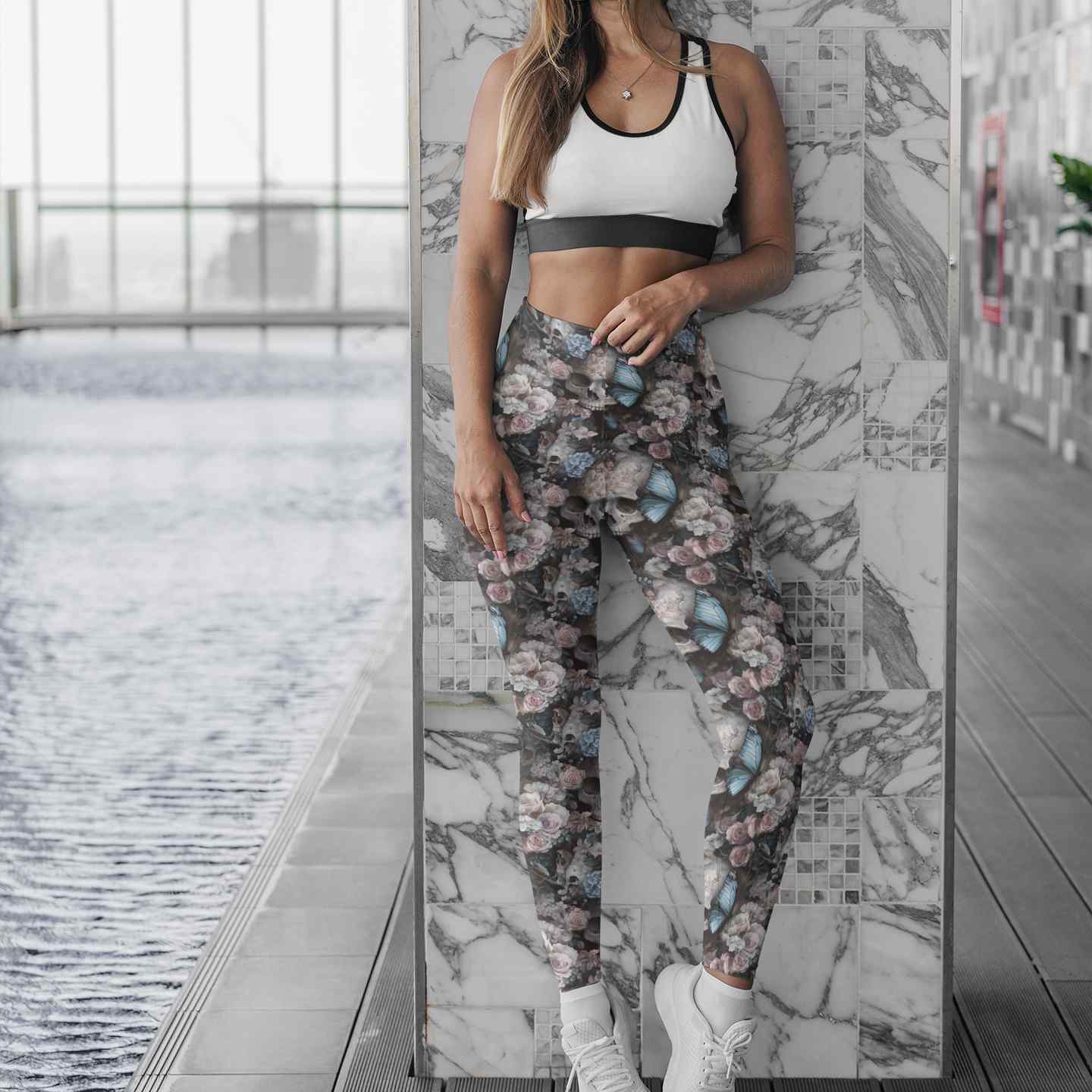 Woman wearing a white sports bra and floral skull and butterfly leggings standing against a marble wall.
