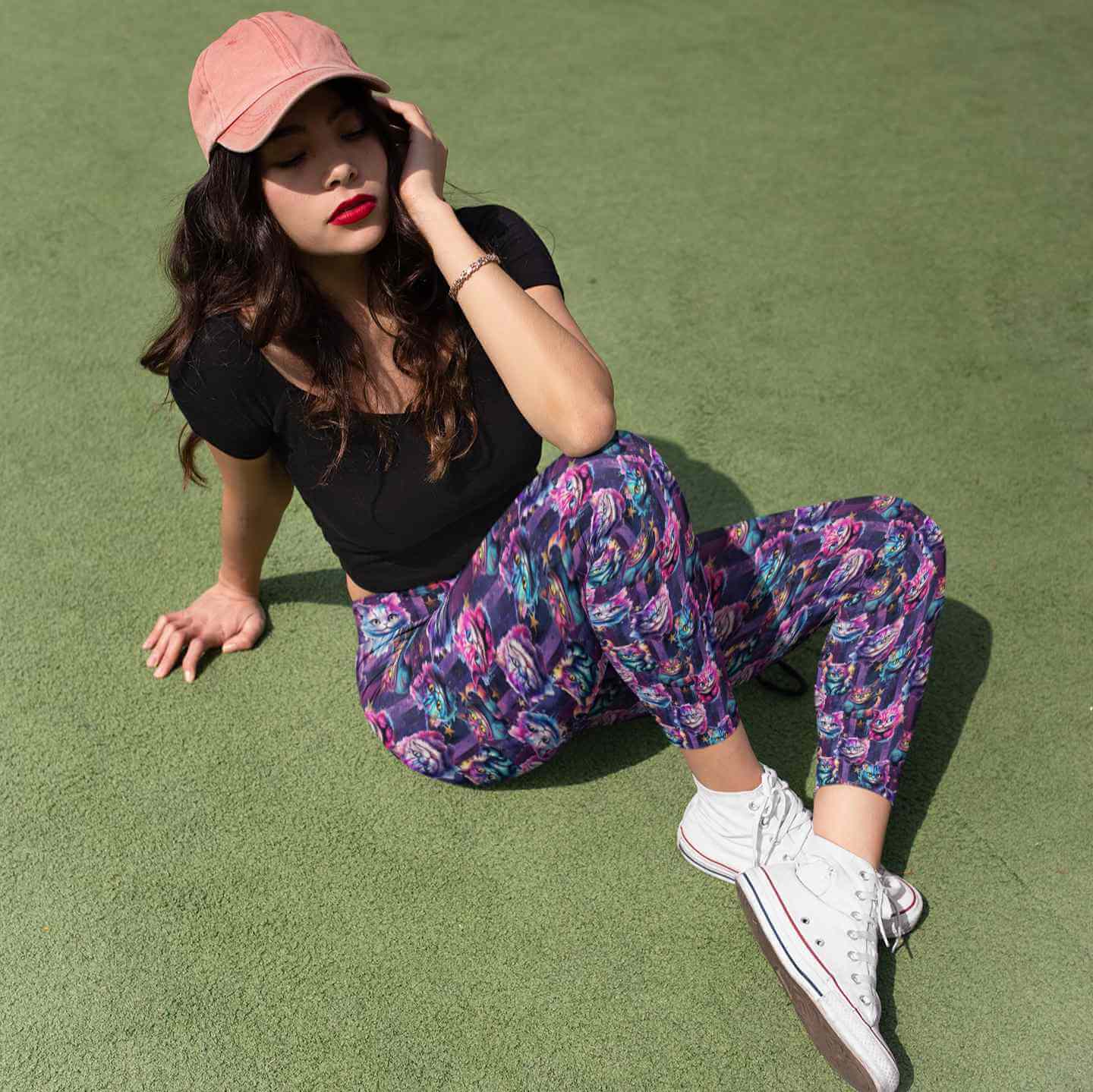 Woman sitting on grass wearing a black top, colorful cat leggings, white sneakers, and a pink cap.