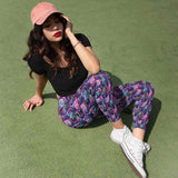 Woman sitting on grass wearing a black top, colorful cat leggings, white sneakers, and a pink cap.