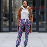 Woman wearing a brown cardigan, white hoodie, Full length jogger pants featuring colourful cosmic cats with bright blue, pink and purple fur, yellow eyes and mischievous smiles standing on a city street.