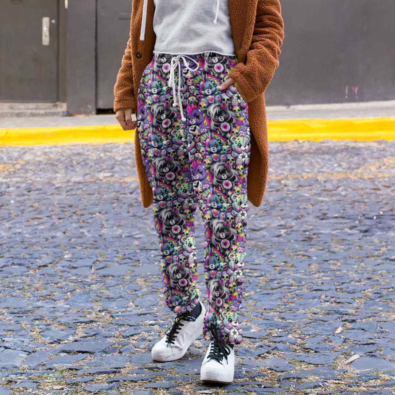 Person wearing Full length jogger pants featuring colourful punk-inspired teddy bears, rainbow skulls, bats, spiderwebs and neon alternative artwork on a black background with bright pink, purple, green and blue accents and a brown coat on a street.
