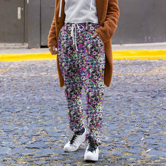 Person wearing Full length jogger pants featuring colourful punk-inspired teddy bears, rainbow skulls, bats, spiderwebs and neon alternative artwork on a black background with bright pink, purple, green and blue accents and a brown coat on a street.