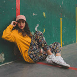 Woman sitting on a basketball court wearing a yellow sweatshirt, floral pants, and white sneakers.