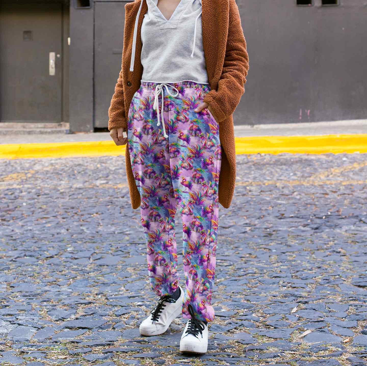 lady on a cobblestone street wearing Full-length jogger pants featuring vibrant pink flamingos dressed in colourful Mardi Gras-style feathers and accessories. The background is a swirling galaxy of purples, pinks and blues with stars and glittery textures.