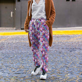 lady on a cobblestone street wearing Full-length jogger pants featuring vibrant pink flamingos dressed in colourful Mardi Gras-style feathers and accessories. The background is a swirling galaxy of purples, pinks and blues with stars and glittery textures.