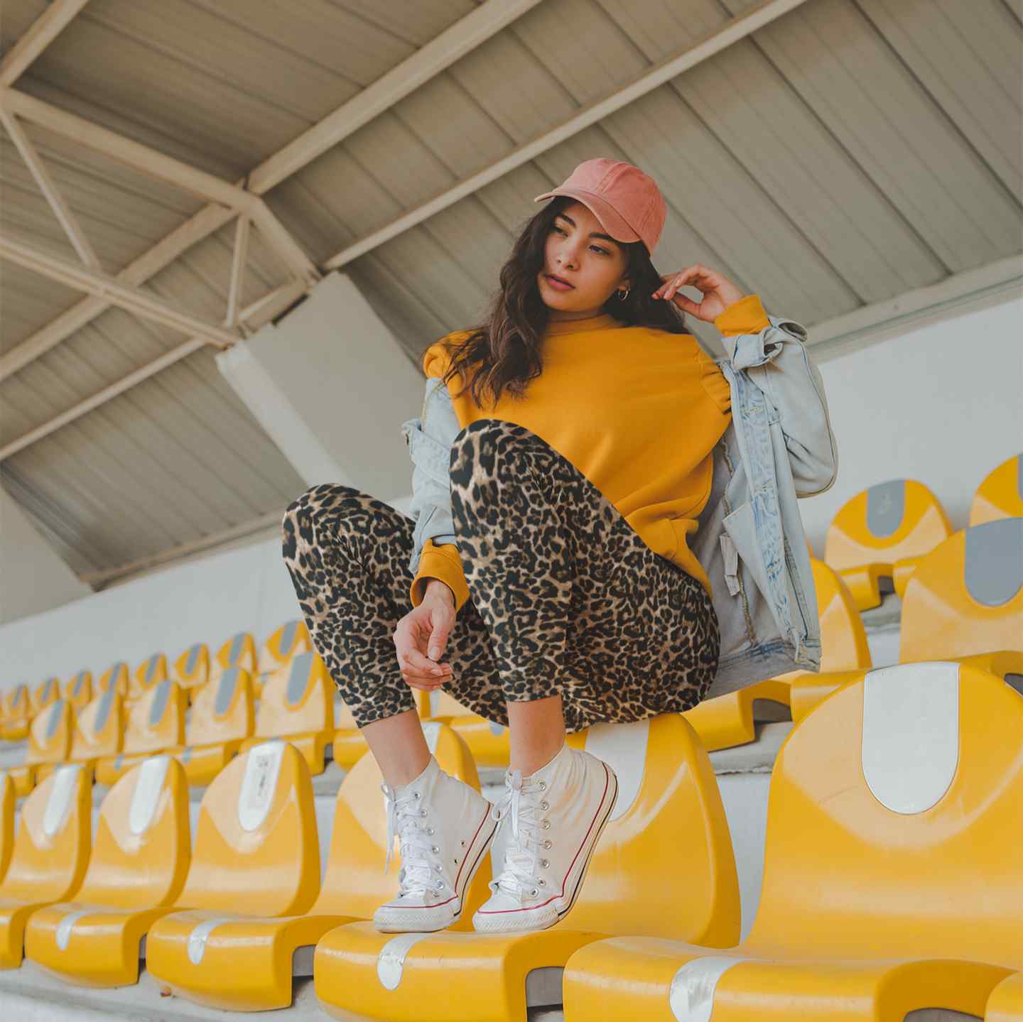 Person sitting on yellow stadium seats wearing a yellow sweater, leopard print pants, and pink cap.